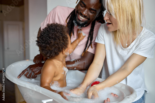 excited black and caucasian family washing daughter child girl in small bathtub, playing with soap and foam, at home, have fun together, laughing. clean skin, skincare, childhood, lifestyle concept