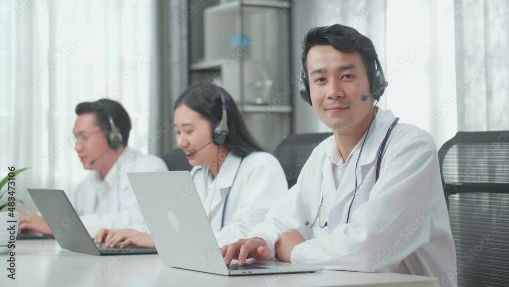 A Man Of Three Asian Doctors With Stethoscope Wearing Headset Working As Call Center Agent Look Up From Computer Then Smile While His Colleagues Are Speaking And Typing During A Call At The Office
A Man Of Three Asian Doctors With Stethoscope Wearing Headset Working As Call Center Agent Look Up From Computer Then Smile While His Colleagues Are Speaking And Typing During A Call At The Office