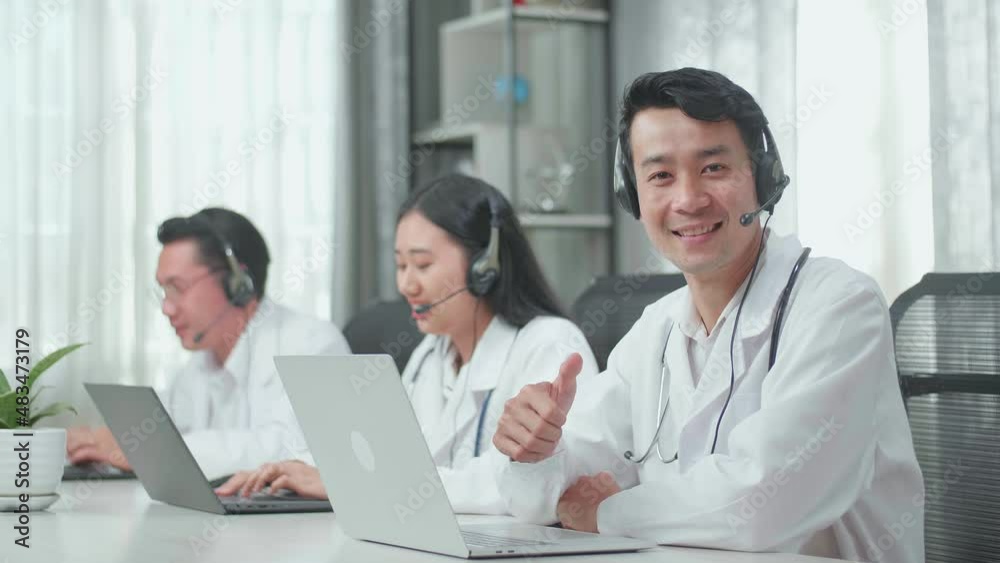 A Man Of Three Asian Doctors With Stethoscope Wearing Headset Working As Call Center Agent Looking Up From Computer Smile Then Thumbs Up While His Colleagues Are Speaking And Typing During A Call At T