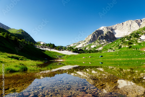 Tent camp on the green flowering shore of a mountain lake surrounded by mountain peaks