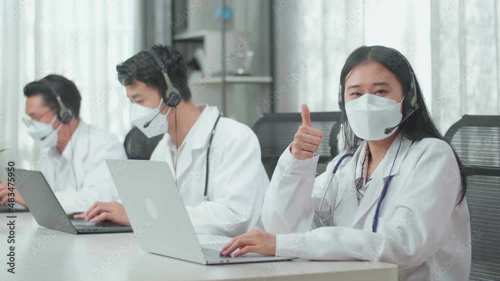 A Woman Of Three Asian Doctors With Stethoscope In Headset And Mask Working As Call Center Agent Look Up From Computer And Thumbs Up While Her Colleagues Are Speaking And Typing During A Call At The O A Woman Of Three Asian Doctors With Stethoscope In Headset And Mask Working As Call Center Agent Look Up From Computer And Thumbs Up While Her Colleagues Are Speaking And Typing During A Call At The O