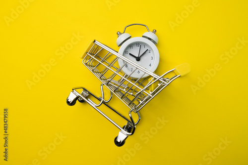White alarm clock in a supermarket trolley on a yellow background. Concept - saving time, buying goods in online stores with delivery, secure, remote shopping during a pandemic. Copy space. Close up.