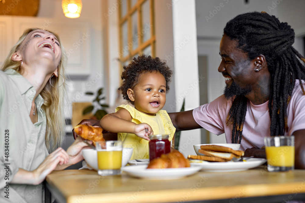 Happy young family with black child girl having breakfast together at table in modern bright cozy kitchen, man woman and kid eating healthy food at home, laughing having talk. friendly family