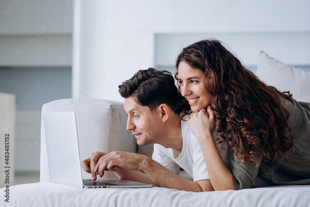 Couple in love lies on the sofa behind a laptop in the living room by the window and makes online purchases or plans a trip
