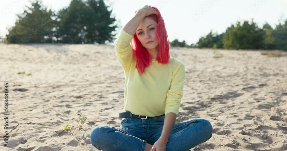 A beautiful serious girl is sitting on the sand outside on the beach, pink hair is blown away by the wind, she blows it out of her face, deep eyes, freckles on face A beautiful serious girl is sitting on the sand outside on the beach, pink hair is blown away by the wind, she blows it out of her face, deep eyes, freckles on face