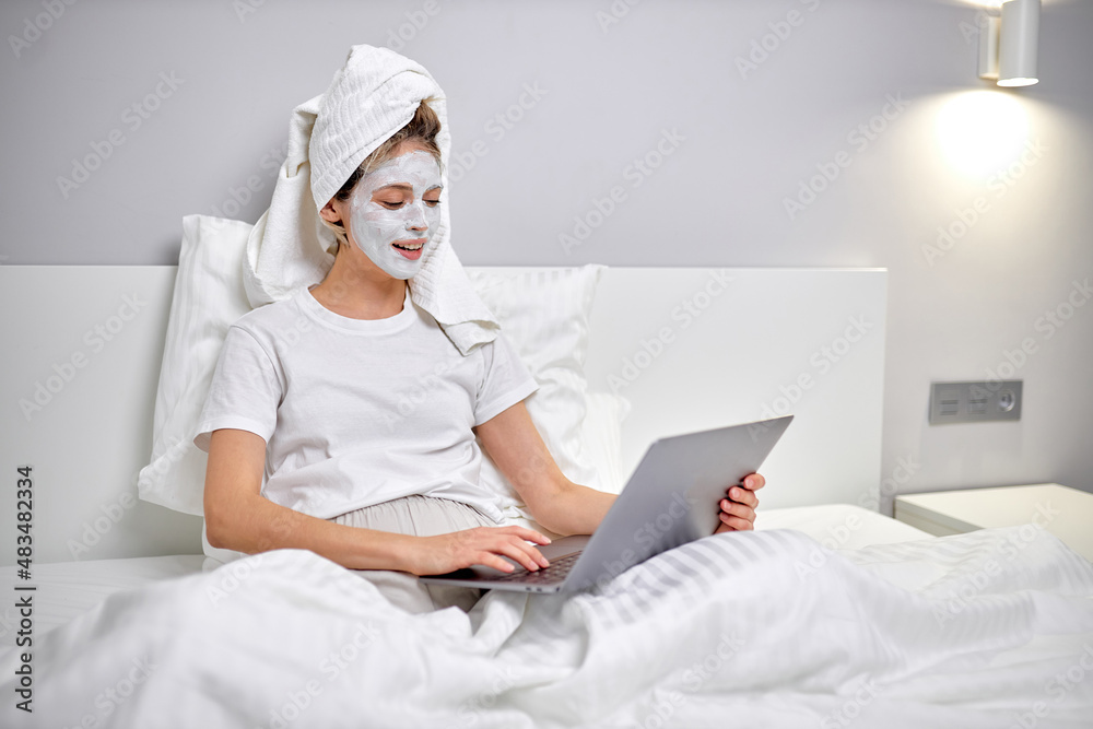 Healthy woman sitting on bed with face beauty mask using laptop and working online. Cozy time at home. Self time and home spa concept. Coronavirus outbreak and self-quarantining concept Healthy woman sitting on bed with face beauty mask using laptop and working online. Cozy time at home. Self time and home spa concept. Coronavirus outbreak and self-quarantining concept
