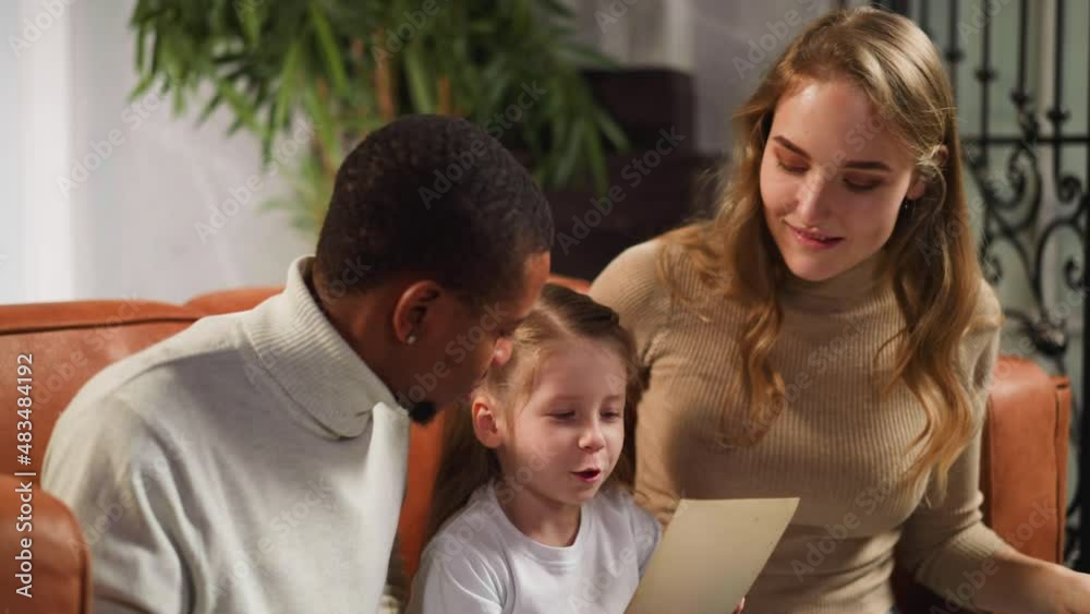 Happy blonde woman turns page of photo album and black father shows picture of grandfather to preschooler girl in living room closeup slow motion Happy blonde woman turns page of photo album and black father shows picture of grandfather to preschooler girl in living room closeup slow motion