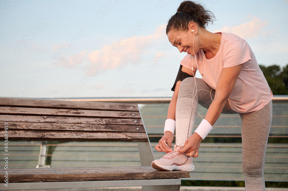 Happy sportswoman with white terry bracelets and smartphone holder smiling while listening to music on headphones, tying sneaker laces, putting foot on wooden bench before morning run. Sportwoman