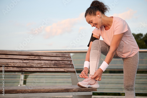 Happy sportswoman with white terry bracelets and smartphone holder smiling while listening to music on headphones  tying sneaker laces  putting foot on wooden bench before morning run. Sportwoman