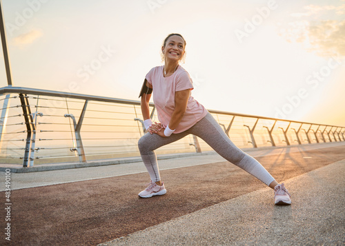 Attractive sporty woman with healthy body doing side lunges, stretching her legs muscles while exercising outdoor on a city bridge at dawn. Sport, fitness, body care and active lifestyle concept