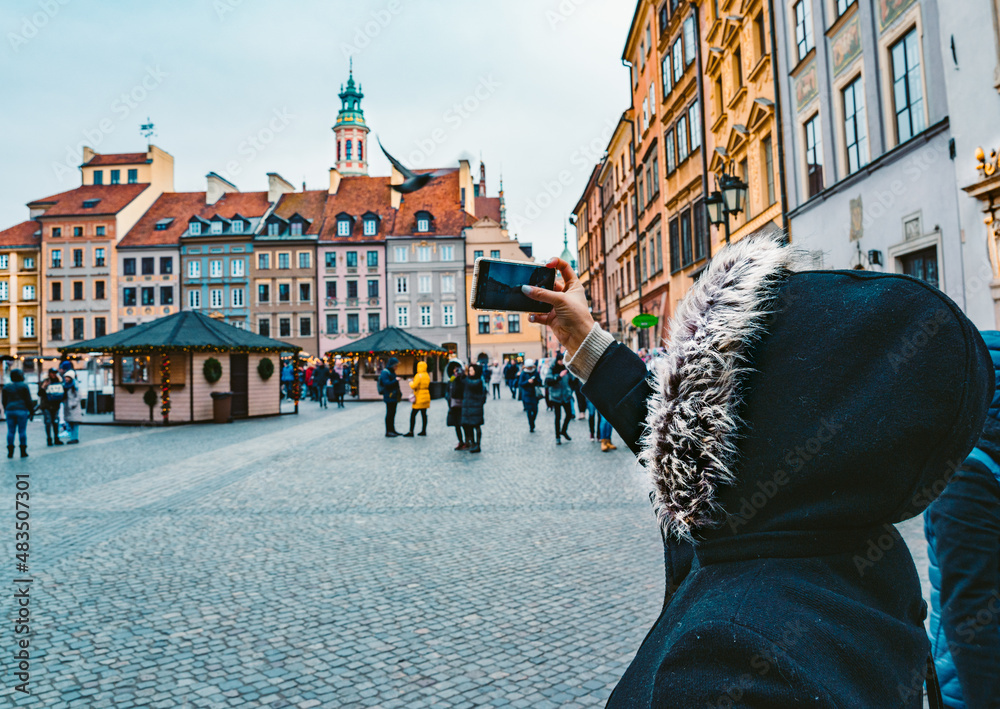 Obraz premium Young Woman in a Fur Hooded Coat Takes a Photo on Phone of Old Town Market Place in Warsaw Poland