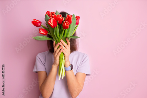 International Women's Day. a happy woman in a purple T-shirt hides her face behind a bouquet of spring tulip flowers that she holds in her hands. Space for text. Very peri #483530720