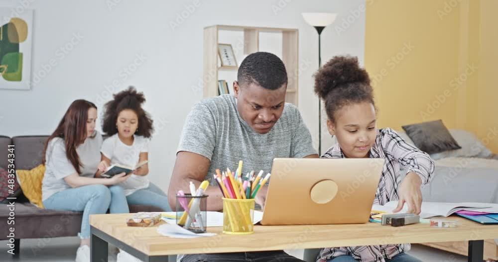 Diverse parents helping kids to do homework. Black father using laptop and helping daughter to do homework assignment near mother and other child sitting on sofa and reading book in living room at Diverse parents helping kids to do homework. Black father using laptop and helping daughter to do homework assignment near mother and other child sitting on sofa and reading book in living room at