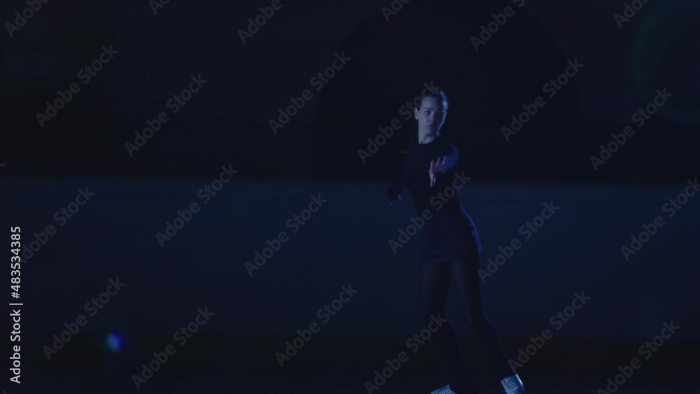 Professional figure skating artist slides and spins on ice arena in rays of blue light. Young woman practicing repertoire of an ice show. Artistic sport, grace, precision and perfection. Slow motion. Professional figure skating artist slides and spins on ice arena in rays of blue light. Young woman practicing repertoire of an ice show. Artistic sport, grace, precision and perfection. Slow motion.
