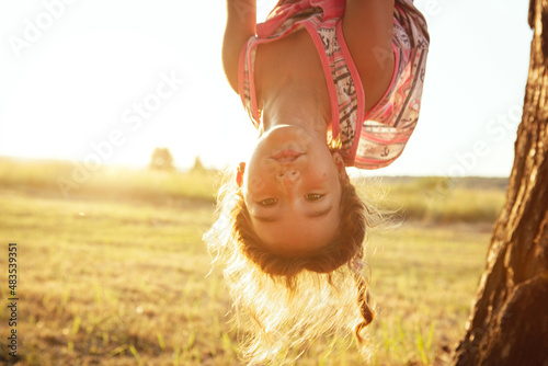 The girl is hanging upside down on a tree in summer in orange sunlight and a light dress. Summer time, heat, childhood. Funny portrait with disheveled hair