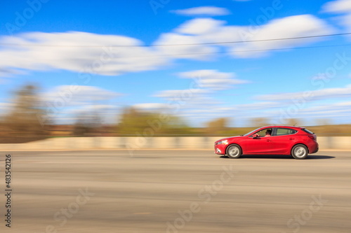 A red car is driving fast on the road on a sunny summer day  the car is in focus  the background is blurred.