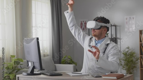 Young male doctor in white gown and VR headset sitting at desk in cabinet and tapping, scrolling with hands in air