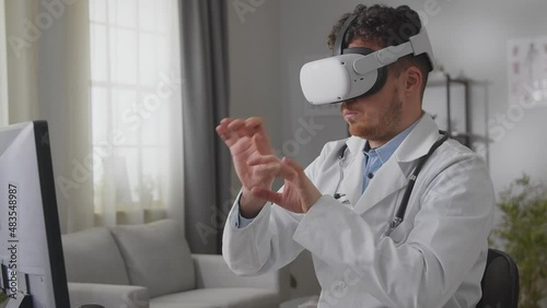 Caucasian male doctor in white gown and VR headset sitting at desk in cabinet and tapping