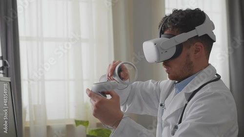 Young adult mane doctor in white gown and VR headset sitting at desk in cabinet and use controller for virtual operation