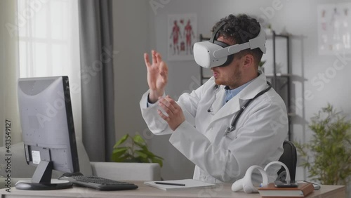 Young male doctor hosts video conference in white VR headset sitting at desk in cabinet