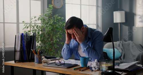 Young tired man working at home office, looking very tired, suffering from splitting headache. Exhausted manager sitting at workplace, trying to cope with anxiety.
