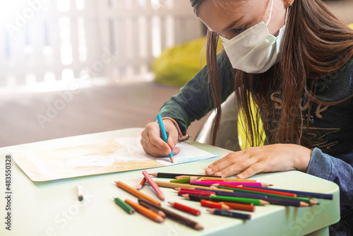A child in a medical mask draws with colored pencils at a table in a bright room. Place for text