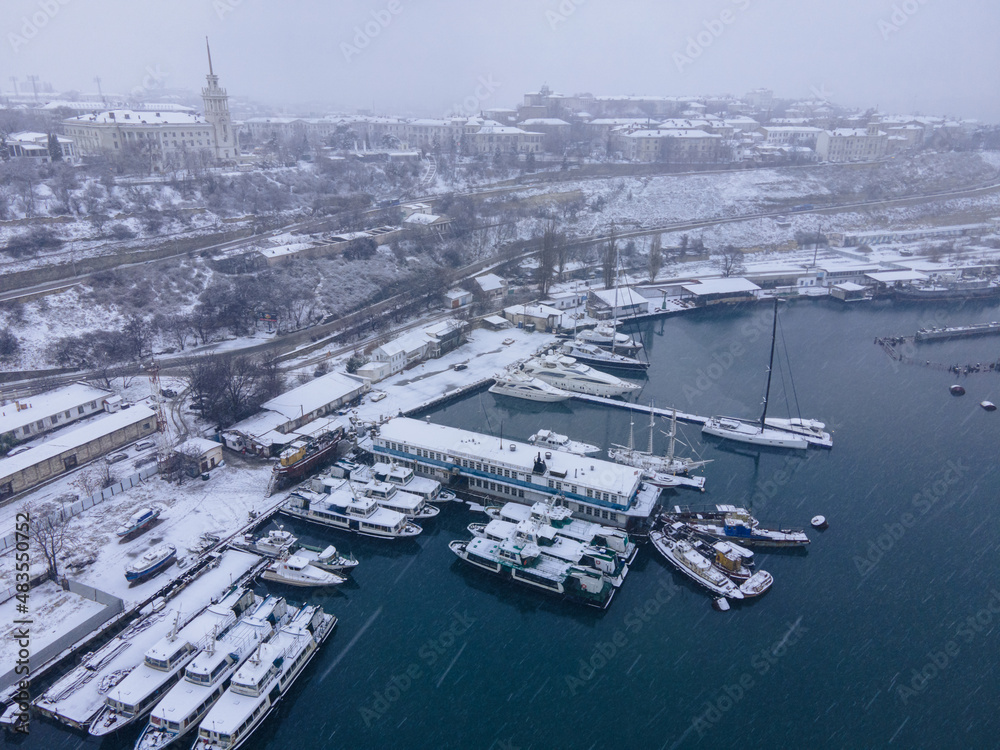 Snow-covered ships in the bay from the air. Old rusty boats in the snow. Winter sea with a snowstorm. Winter fairy tale in the city from the air.