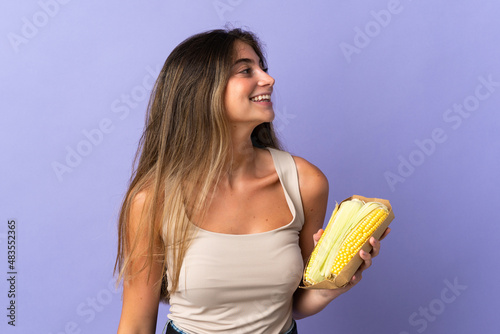 Young woman holding corn isolated on purple background looking up while smiling