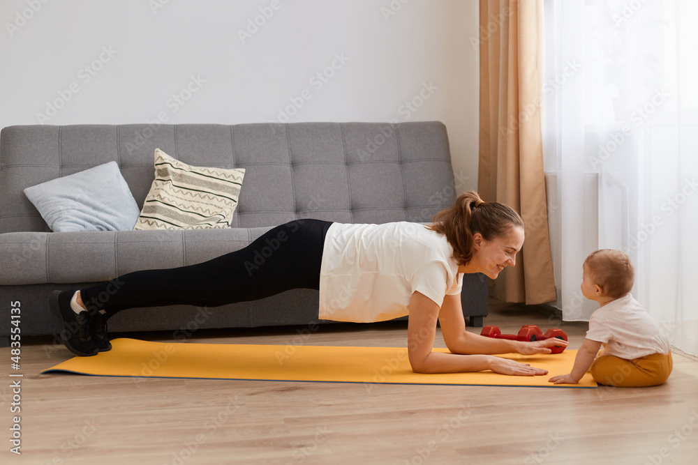Side view of young woman exercising, standing in plank position to train her abs, arms endurance and strength on yoga mat at home in living room with her infant baby daughter. Side view of young woman exercising, standing in plank position to train her abs, arms endurance and strength on yoga mat at home in living room with her infant baby daughter.