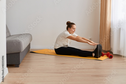 Full length profile portrait of beautiful athletic young woman practicing yoga at home, sitting on mat and stretching her legs, doing sport exercises for her health, fitness and lifestyle.