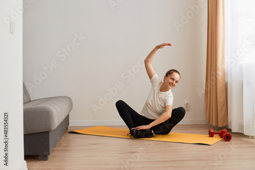 Full length portrait of young healthy beautiful woman in T-shirt and leggings practicing yoga at home sitting in lotus pose on yoga mat and tiling her body to side, stretching.