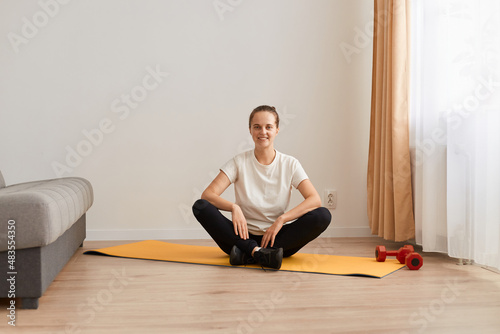 Portrait of attractive sporty woman sitting in lotus pose on mat, wearing white t shirt and black leggins, looking at camera with happy smile, meditating and training at home in living room.