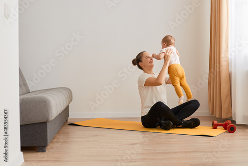 Portrait of athletic woman sitting in lotus pose on mat, wearing white t shirt and black leggins, posing in living room and throwing her baby daughter to the air while having rest, healthy lifestyle.