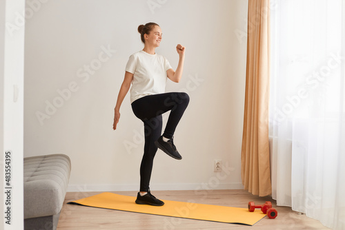 Full length portrait of slim athletic woman wearing white t shirt and black leggins doing sport exercises at home on yoga mat, walking in place, marching, sport, fitness at home.