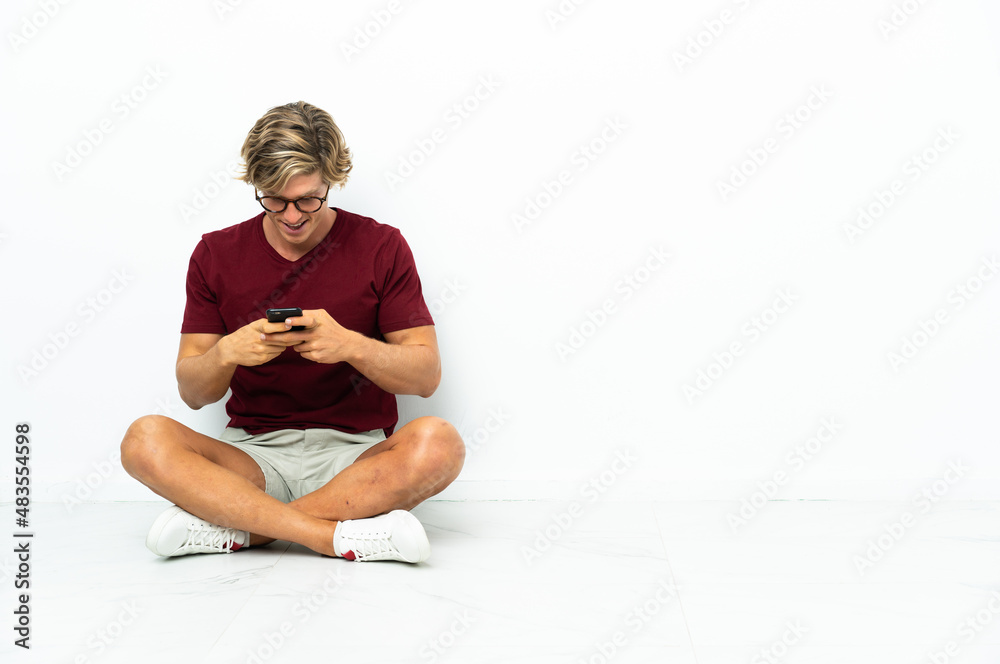 Fototapeta premium Young English man sitting on the floor sending a message with the mobile