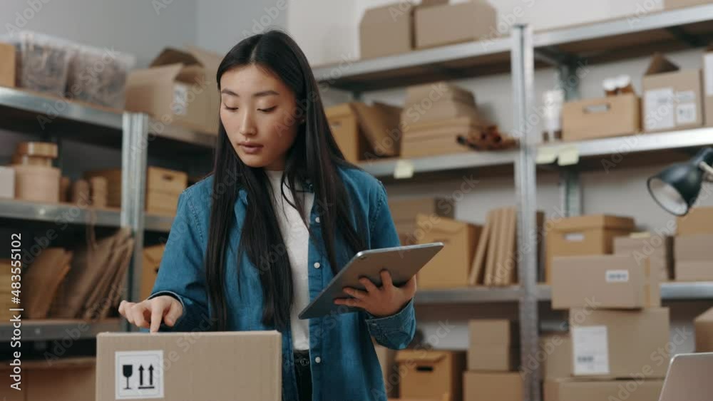 Calm asian woman using tablet computer while browsing something on it and preparing sending parcel to the client. Girl working at the warehouse. Small business concept. Calm asian woman using tablet computer while browsing something on it and preparing sending parcel to the client. Girl working at the warehouse. Small business concept.