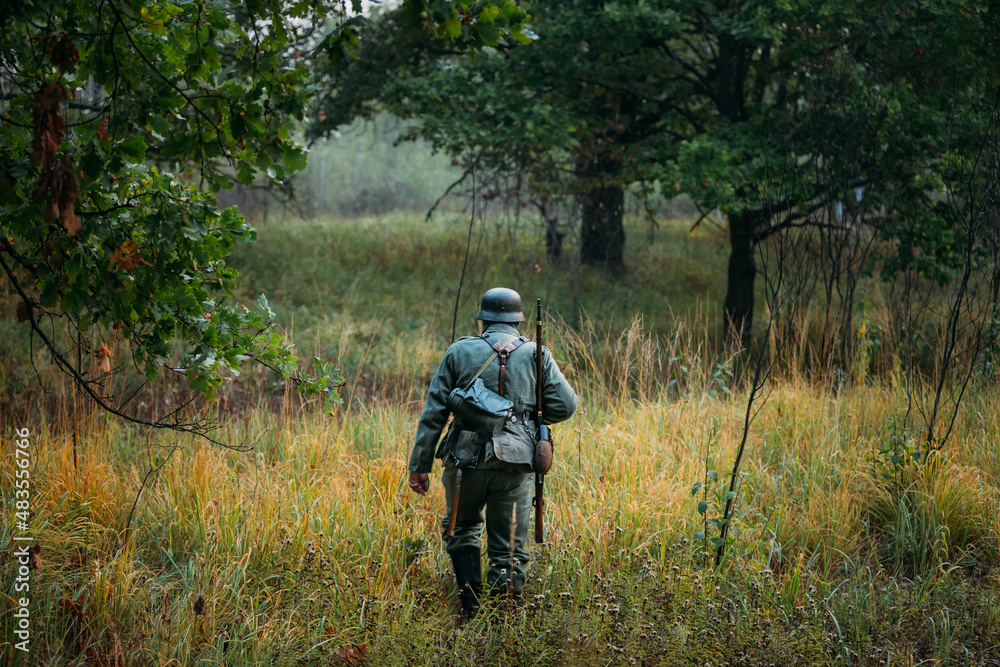 Single Re-enactor Dressed As German Wehrmacht Infantry Soldier In World War II Walking In Patrol Through Autumn Forest. WWII WW2 Times. Single Re-enactor Dressed As German Wehrmacht Infantry Soldier In World War II Walking In Patrol Through Autumn Forest. WWII WW2 Times.