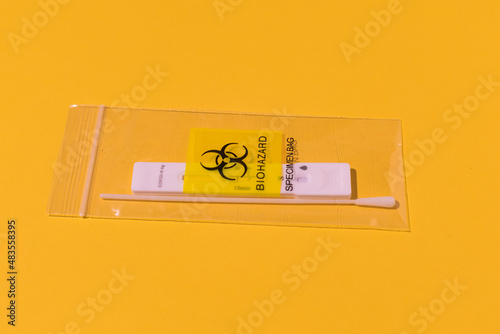 Plastic sample bag with the biohazard logo on it, with antigen test and swab used inside, on a yellow background. Waste, contamination, test, covid-19, virus, omicron, delta and ihu concept. photo