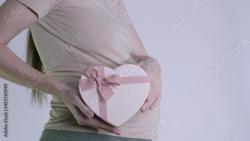 a pregnant woman shows a pink gift box in the shape of a heart on a belly background. on a white background