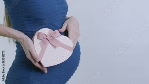 A pregnant woman in a blue dress receives a gift on Valentine's Day or March 8. Pink heart-shaped box with a bow on the background of the belly. video in slow motion.