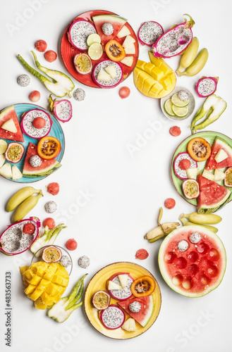 Exotic fruits on colorful plates and white table. Cirle frame. Watermelon, ananas, pitaya, banana, passion fruits, lime and mango. Healthy and delicious tropical fruits. Top view with copy space.