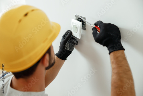 Professional electrician in uniform using screwdriver while installing electrical socket outlet after renovation work