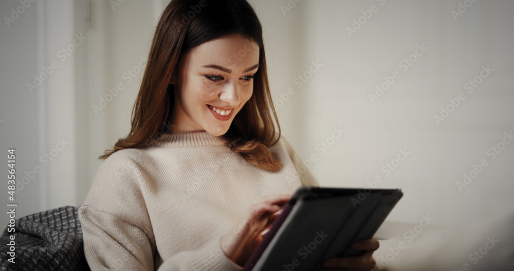 Charming young woman enjoys using new app on tablet, working online at cozy home atmosphere. Smiling lady using social networks to rest or chat with friends. Charming young woman enjoys using new app on tablet, working online at cozy home atmosphere. Smiling lady using social networks to rest or chat with friends.