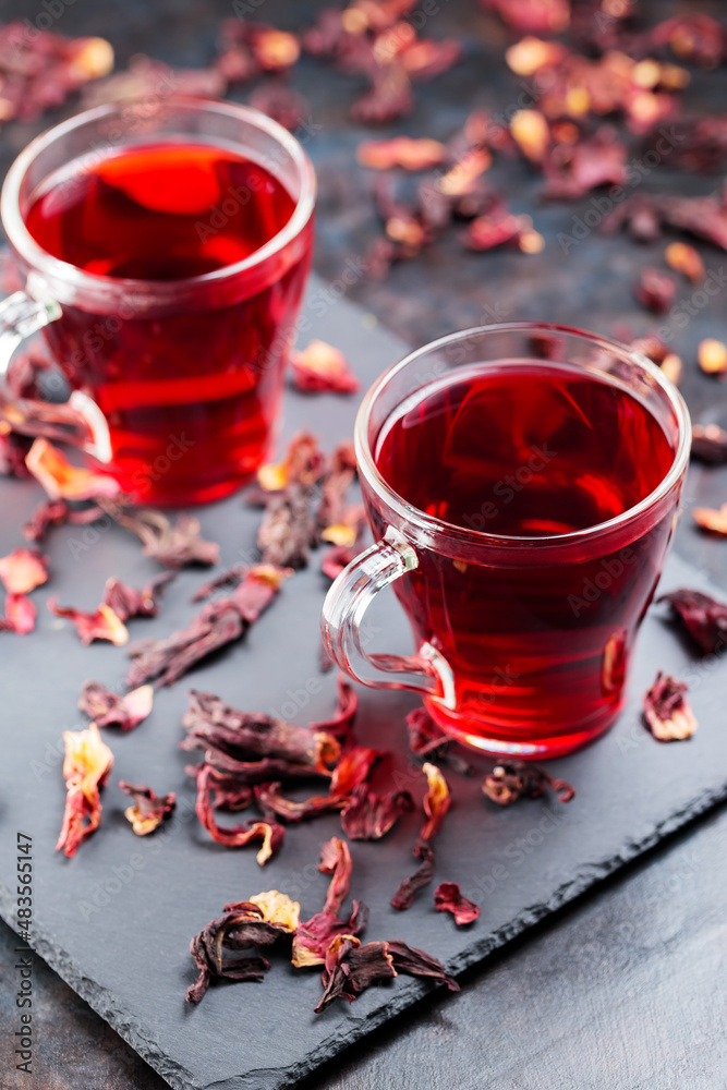 Hibiscus tea in glass mug on a dark background. Cup of red hibiscus tea and dry hibiscus petals on a slate board Hibiscus tea in glass mug on a dark background. Cup of red hibiscus tea and dry hibiscus petals on a slate board
