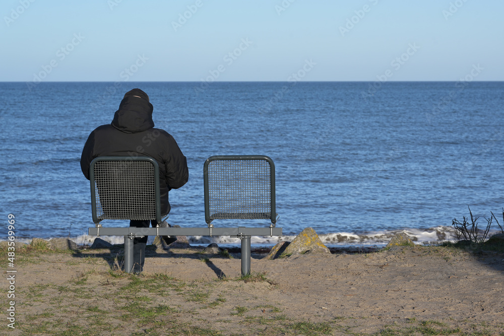 Single man in a dark warm jacket from behind sitting next to an empty chair on the sea shore looking over the water to the horizon, recreation or mourning for  loss of a loved person, copy space