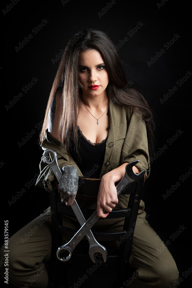 Piękna dziewczyna z tatuażami przebrana w strój mechanika, siedzi na krześle i trzyma bardzo duże klucze. Zdjęcie zrobione w studio na czarnym tle, oświetlenie z 2 lamp. - obrazy, fototapety, plakaty 