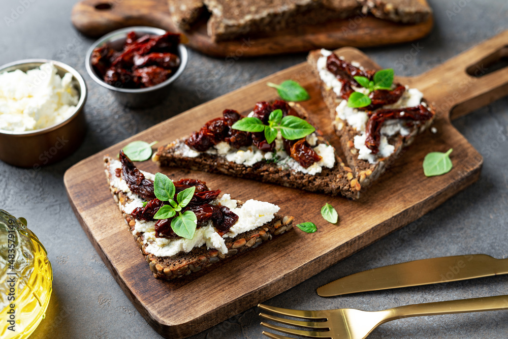 Bruschetta or whole grain bread sandwich with cream cheese and sun-dried tomatoes. Crostini on a wooden serving board on a dark kitchen table closeup