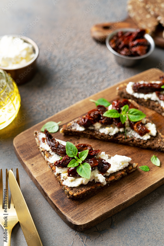 Bruschetta or whole grain bread sandwich with cream cheese and sun-dried tomatoes. Crostini on a wooden serving board on a dark kitchen table closeup