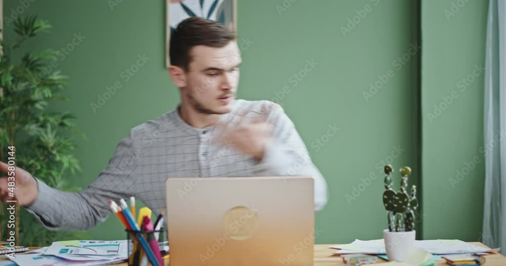 Young brunet man with stubble sitting in office feels stressed and can not find right work papers in mess on table with laptop closeup