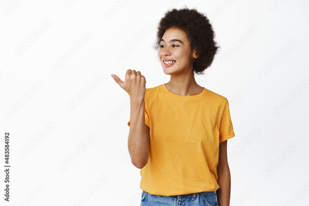 Happy black woman pointing and looking left with pleased smile, standing in yellow t-shirt over white background Happy black woman pointing and looking left with pleased smile, standing in yellow t-shirt over white background